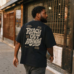 Man seen from behind wearing a black t-shirt with large cream graphic text reading REACH OUT ANYWAY, walking down a city street