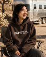 Woman laughing outdoors in a desert campsite wearing a brown hoodie with 'You Are Bad For My Mental Health' text